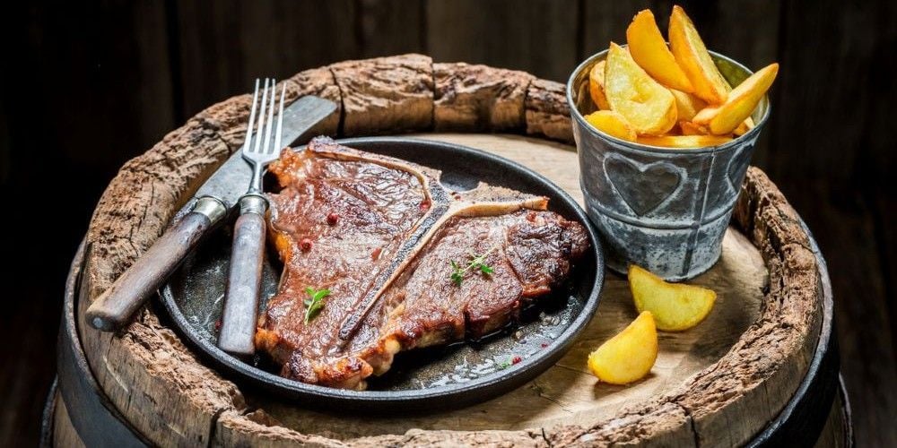 Grilled T-bone steak on a cast iron plate with cutlery and a bucket of potato wedges on a rustic wooden barrel